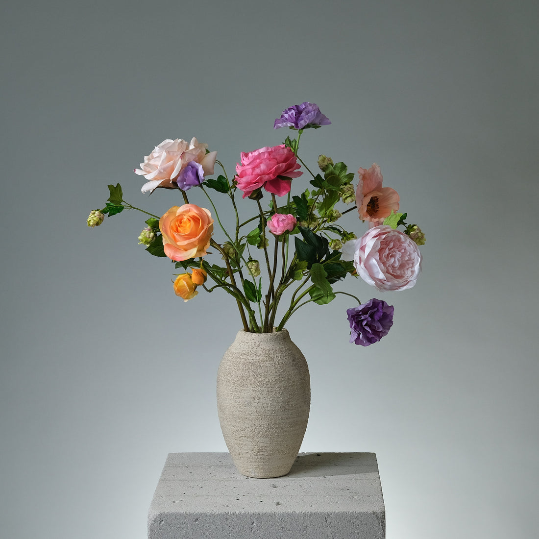Bukett i konstblommor med ljusrosa pion, rosa vallmo, gula och rosa rosor, lavendelfärgad pensé, mörkrosa ranunkel och gröna humleblad i en romantisk och livfull komposition.- FLORISSMA